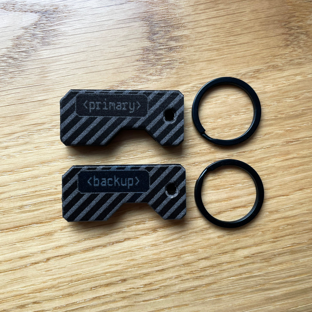 Czarny zestaw dwóch ochronnych etui na klucze YubiKey z grawerem „primary” oraz „backup”. Widoczny klucz YubiKey 5C NFC oraz YubiKey 5 NFC w wersji USB-A wysunięte z obudów. Idealne rozwiązanie do odróżnienia klucza głównego od zapasowego. Czarne kółka do kluczy.